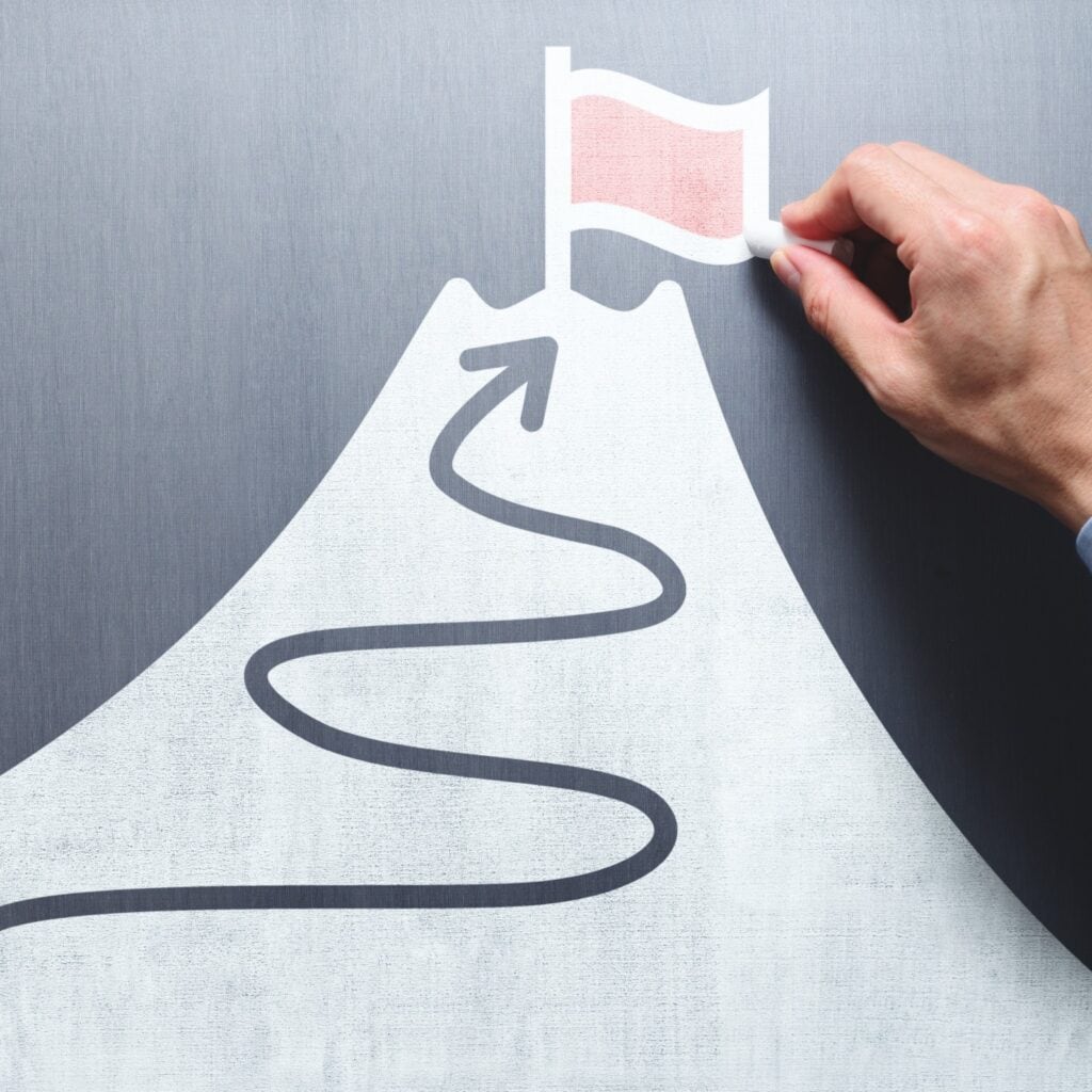 Business goal and success concept. Businessman drawing flag and mountain on chalkboard.
